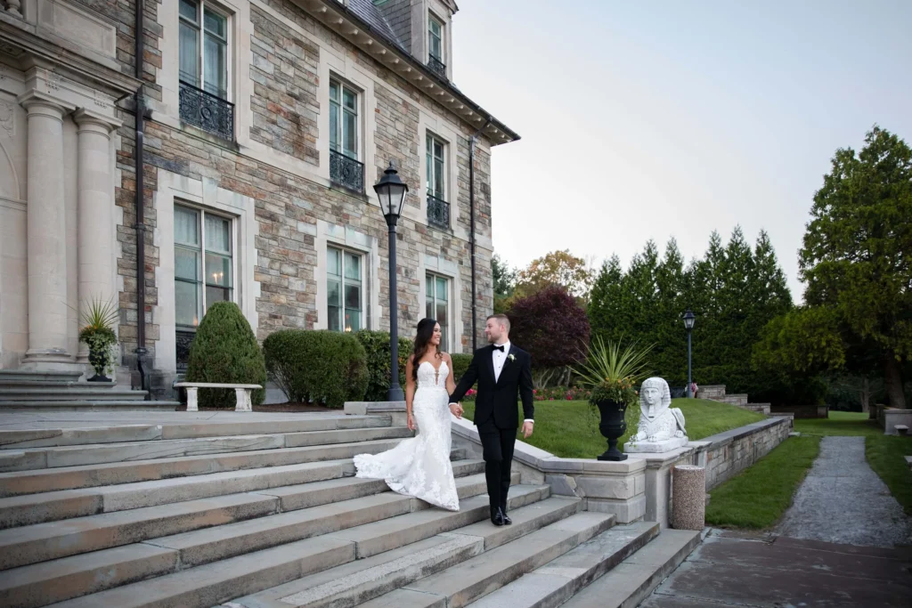 Aldrich Mansion | Kelsey Sheehan Photography Rhode Island Wedding Photographer | A couple in formal attire walks down grand stone steps outside a stately mansion. The scene is elegant, with manicured hedges and serene ambiance.