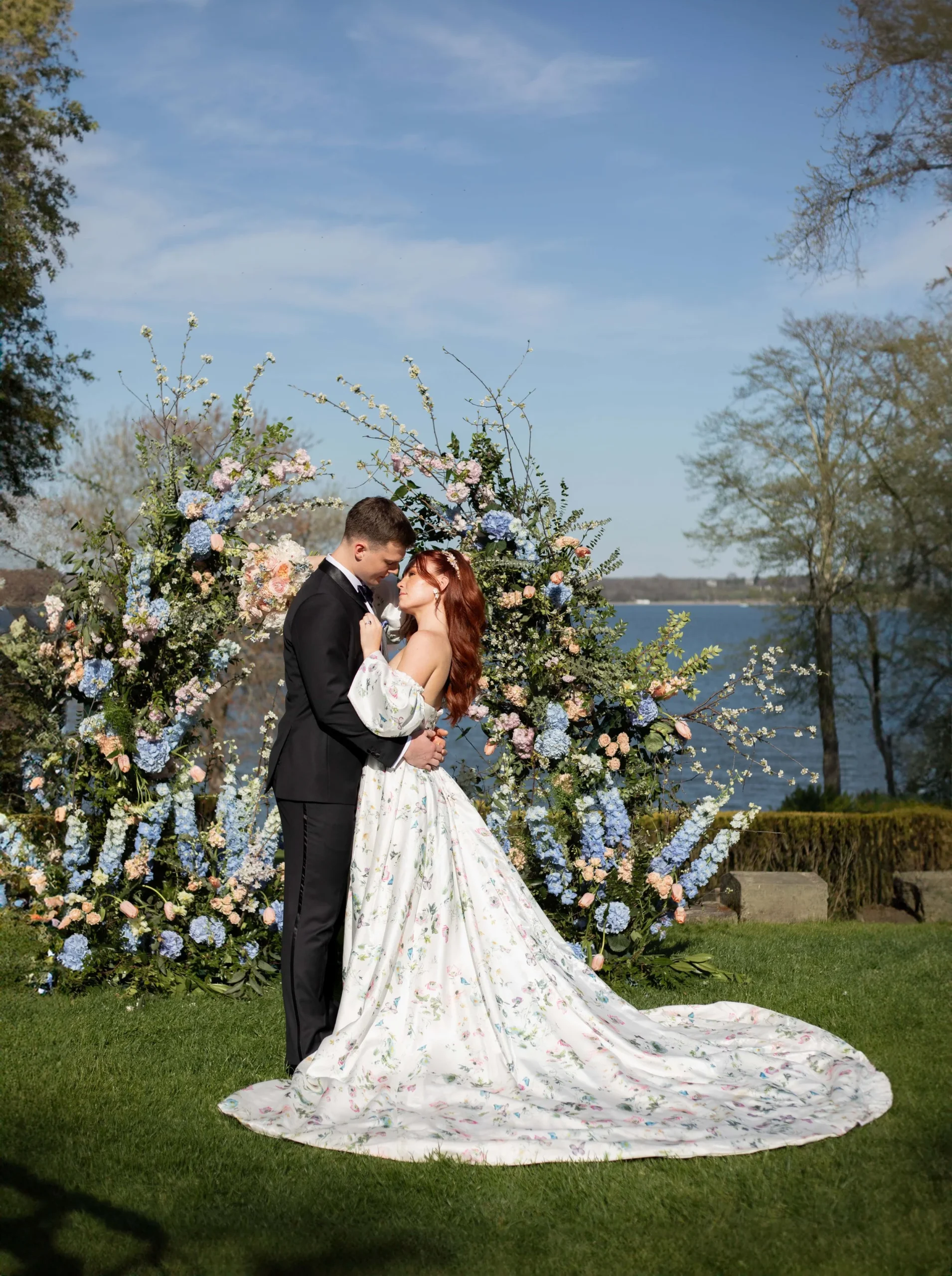 Glen Manor House | Kelsey Sheehan Photography Rhode Island Wedding Photographer | Bride and groom embrace under a lush floral arch by a lakeside. She wears a flowy floral gown; he is in a black suit. The scene is romantic and serene.