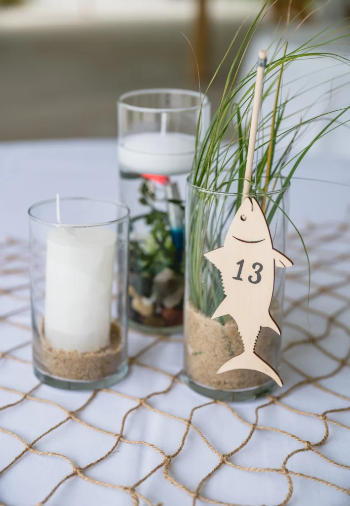 Bristol Harbor Inn | Kelsey Sheehan Photography Rhode Island Wedding Photographer | Three glass vases atop a nautical net on a table. One holds a candle on sand, another with greenery and a wooden fish marked "13". The tone is coastal and serene.