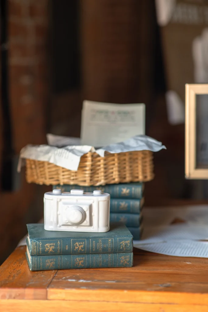 Boston Public Library Engagement Photos Session | Kelsey Sheehan Photography Rhode Island Wedding Photographer | A decorative white camera sits on stacked green books on a wooden table. A wicker basket with papers is in the background, giving a vintage feel.
