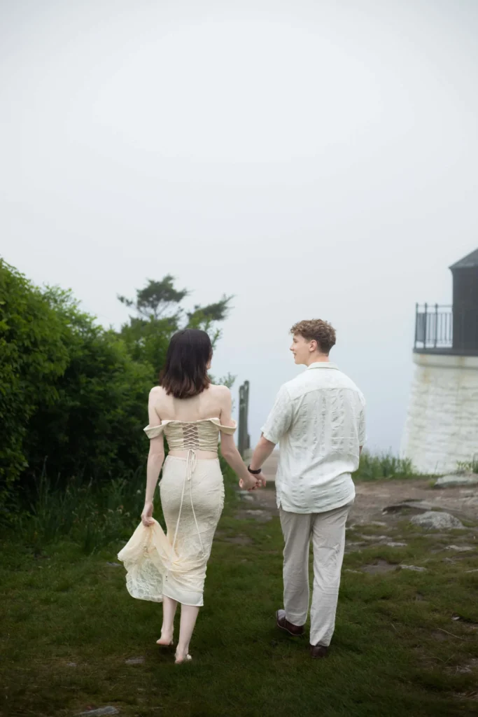 Castle Hill Inn | Kelsey Sheehan Photography Rhode Island Wedding Photographer | A couple walks hand in hand on a foggy day, the woman in an off-shoulder dress and the man in a casual shirt. They approach a lighthouse.