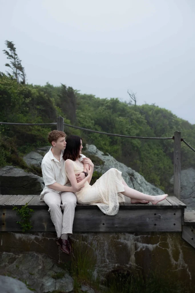 Castle Hill Inn | Kelsey Sheehan Photography Rhode Island Wedding Photographer | A couple sits on a wooden platform overlooking a misty, lush green landscape. They appear relaxed and contemplative, creating a serene and intimate mood.