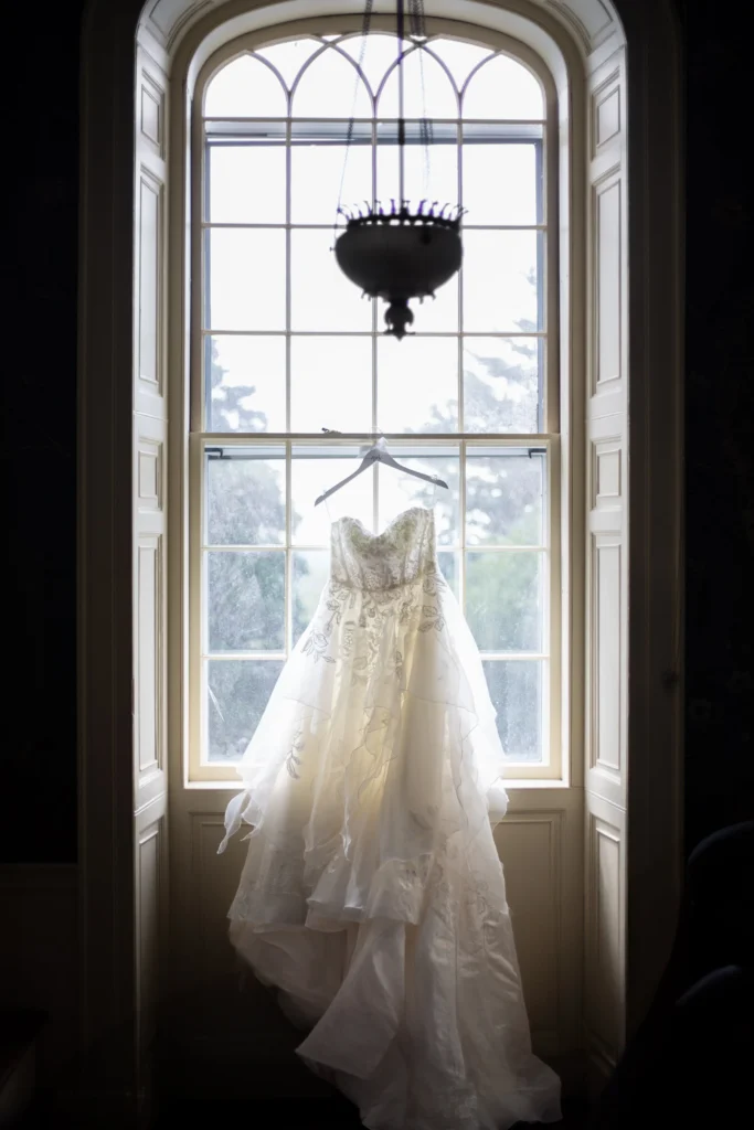 Rosecliff Mansion | Kelsey Sheehan Photography Rhode Island Wedding Photographer | A delicate white wedding dress hangs on a hanger in front of a large arched window, softly backlit, creating a romantic and serene atmosphere.