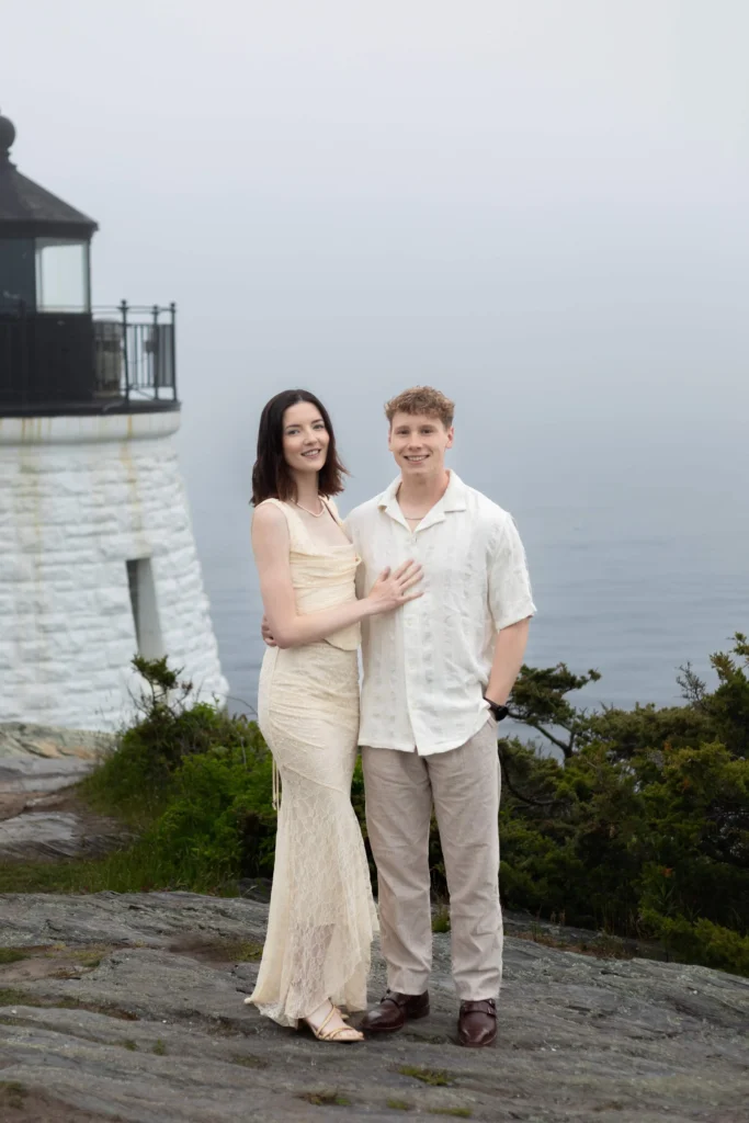Castle Hill Inn | Kelsey Sheehan Photography Rhode Island Wedding Photographer | A smiling couple stands together on a rocky terrain near a white lighthouse, with the ocean in the background. The atmosphere is calm and overcast.