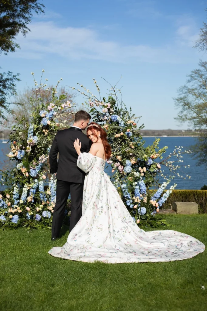 Castle Wedding Venues in New England | Kelsey Sheehan Photography Rhode Island Wedding Photographer | A couple stands by a lake, surrounded by a large floral arch in pastel hues. The woman wears a floral dress, exuding romance against the blue sky backdrop.