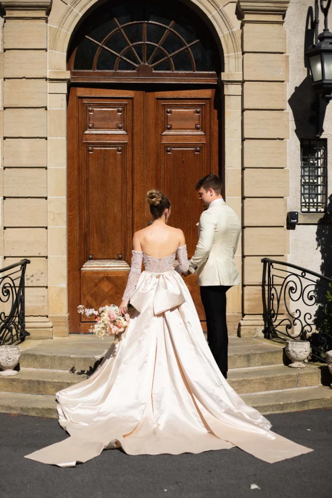 Glen Manor House | Kelsey Sheehan Photography Rhode Island Wedding Photographer | A bride in an elegant, flowing gown and a groom in a light suit stand hand in hand, facing a grand wooden door with an arched frame, conveying romance.