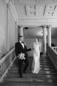 Groom leading his bride down the back staircase at Aldrich Mansion before the ceremony. Bride is laughing and wearing a sleeveless satin dress with a matching shawl. He is wearing a black tux and bowtie and holding her bouquet. 