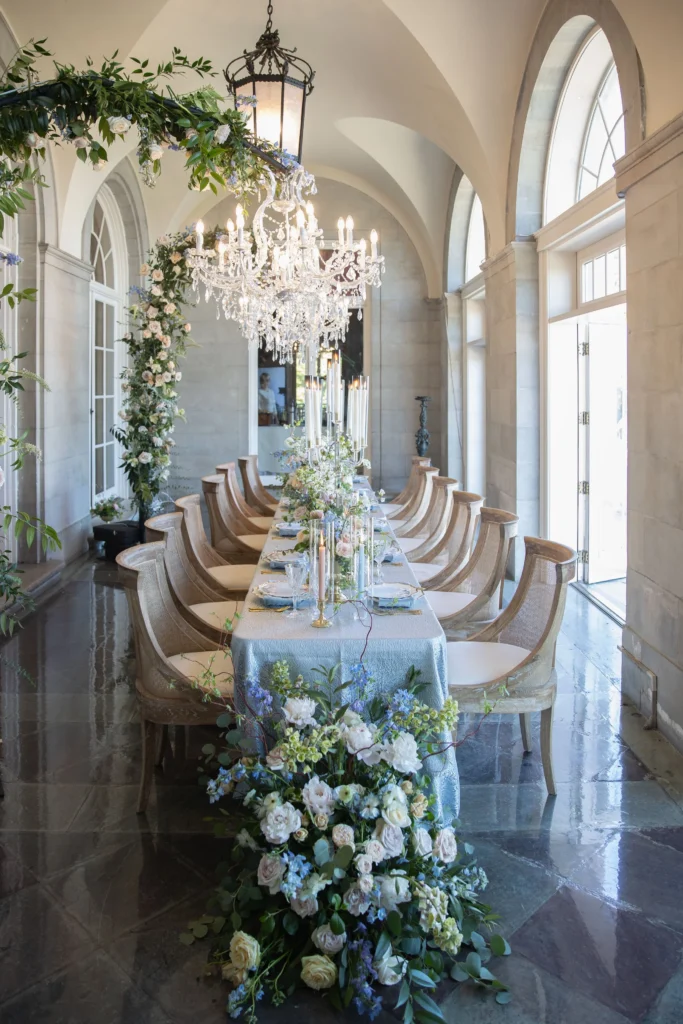 Castle Wedding Venues in New England | Kelsey Sheehan Photography Rhode Island Wedding Photographer | Elegant dining setup with a long table adorned with pastel flowers and candles under a grand chandelier. Arched windows and floral decor create a lavish ambiance.