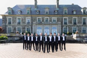 Groom and his groomsmen standing on the back patio of the mansion. All of the guys are wearing black tuxes and bowties. 
