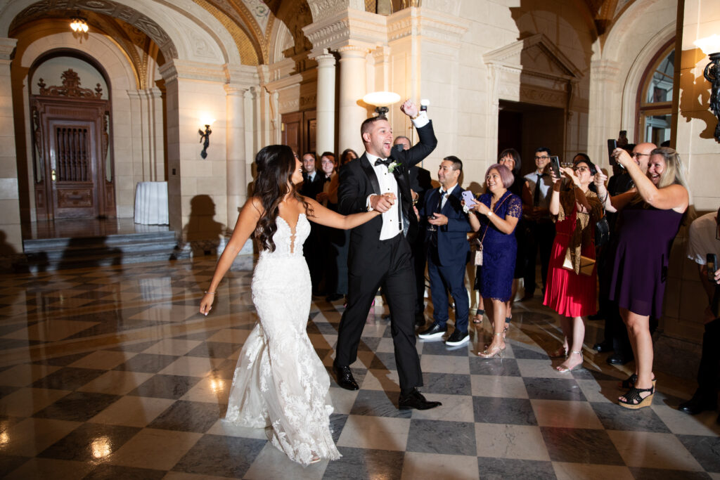 Aldrich Mansion | Kelsey Sheehan Photography Rhode Island Wedding Photographer | Bride and groom joyfully enter a grand hall with arched ceilings and marble floors. Guests cheer and take photos, capturing the celebratory atmosphere.