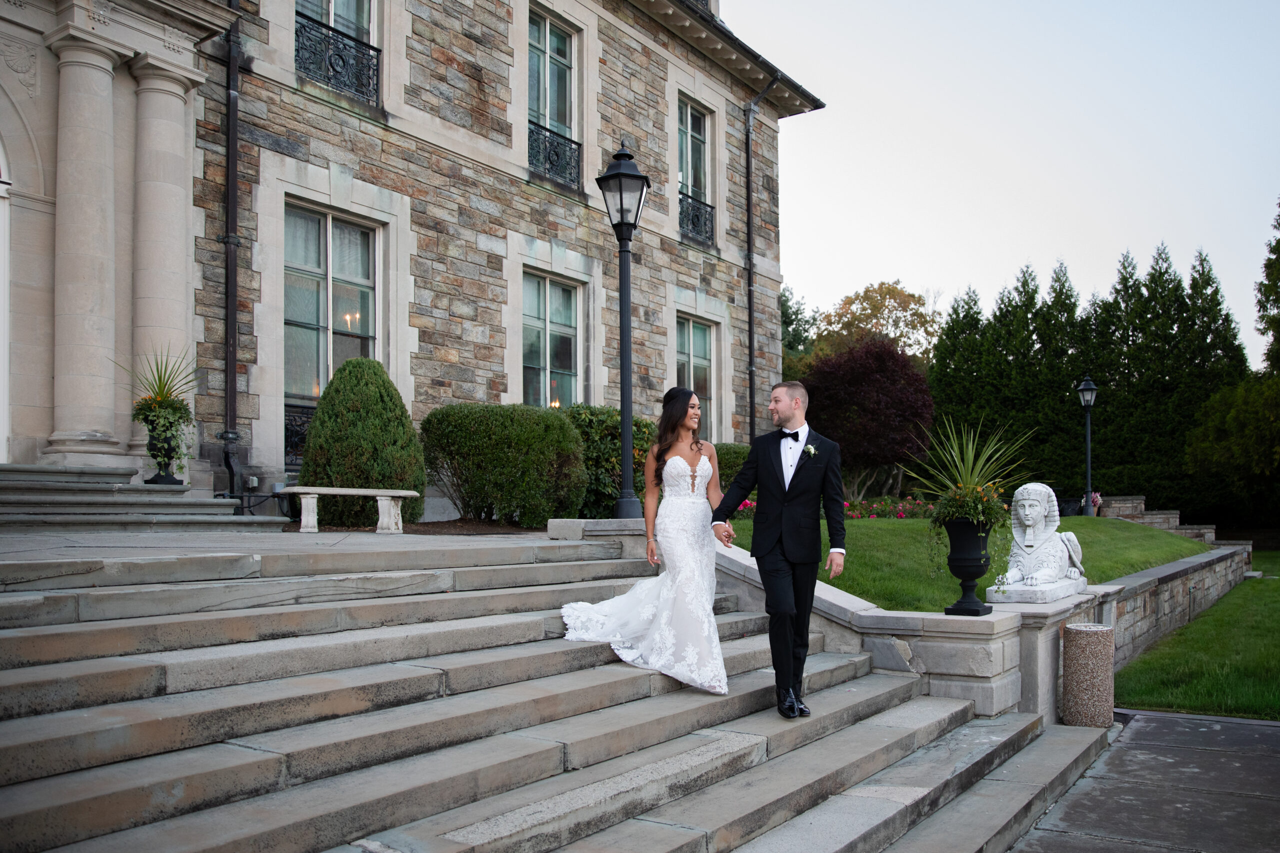 Aldrich Mansion | Kelsey Sheehan Photography Rhode Island Wedding Photographer | A couple in formal attire walks down grand stone steps outside a stately mansion. The scene is elegant, with manicured hedges and serene ambiance.
