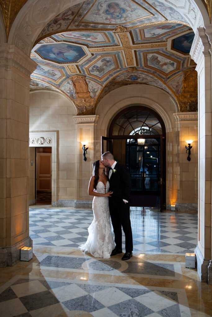 Castle Wedding Venues in New England | Kelsey Sheehan Photography Rhode Island Wedding Photographer | A couple in wedding attire shares a tender moment in an ornate hall with an arched ceiling, detailed murals, and elegant lighting. Romantic ambiance.