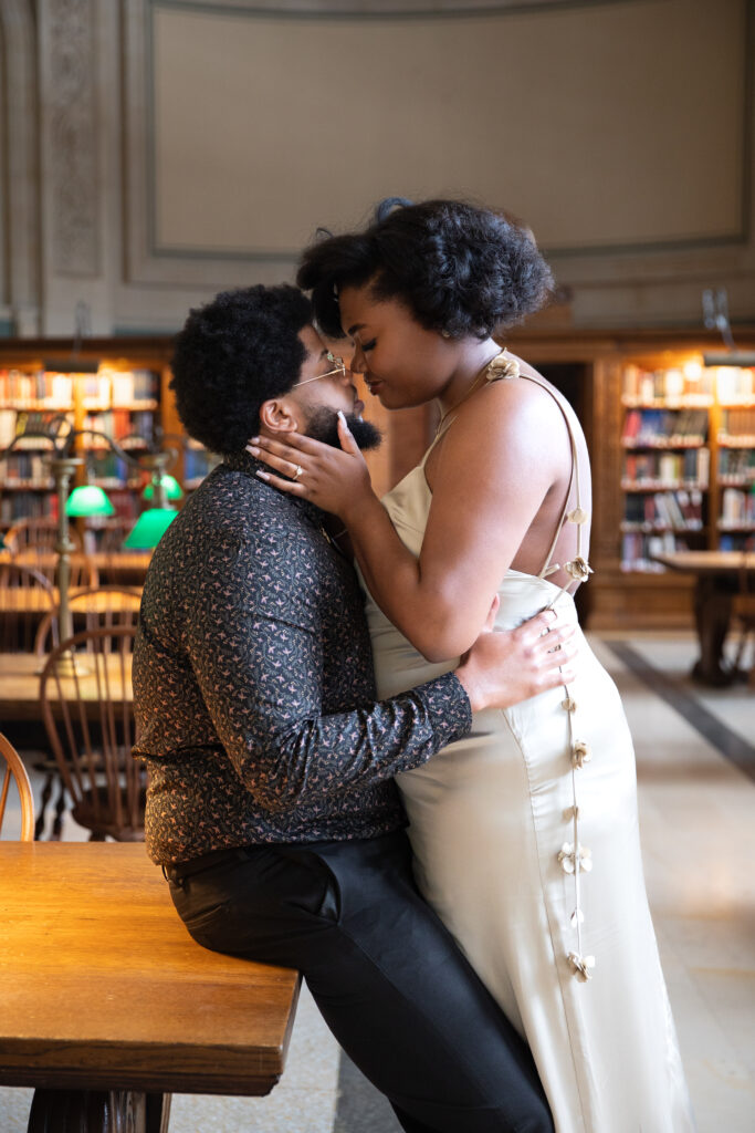Boston Public Library Engagement Photos Session | Kelsey Sheehan Photography Rhode Island Wedding Photographer | A couple shares an intimate moment in a library. The woman in a white dress sits on the man's lap. Warm lighting and bookshelves add a cozy feel.