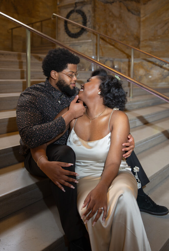 Boston Public Library Engagement Photos Session | Kelsey Sheehan Photography Rhode Island Wedding Photographer | A couple sits on marble stairs in an embrace. The man gently touches the woman's chin, both smiling warmly at each other, conveying romance and closeness.