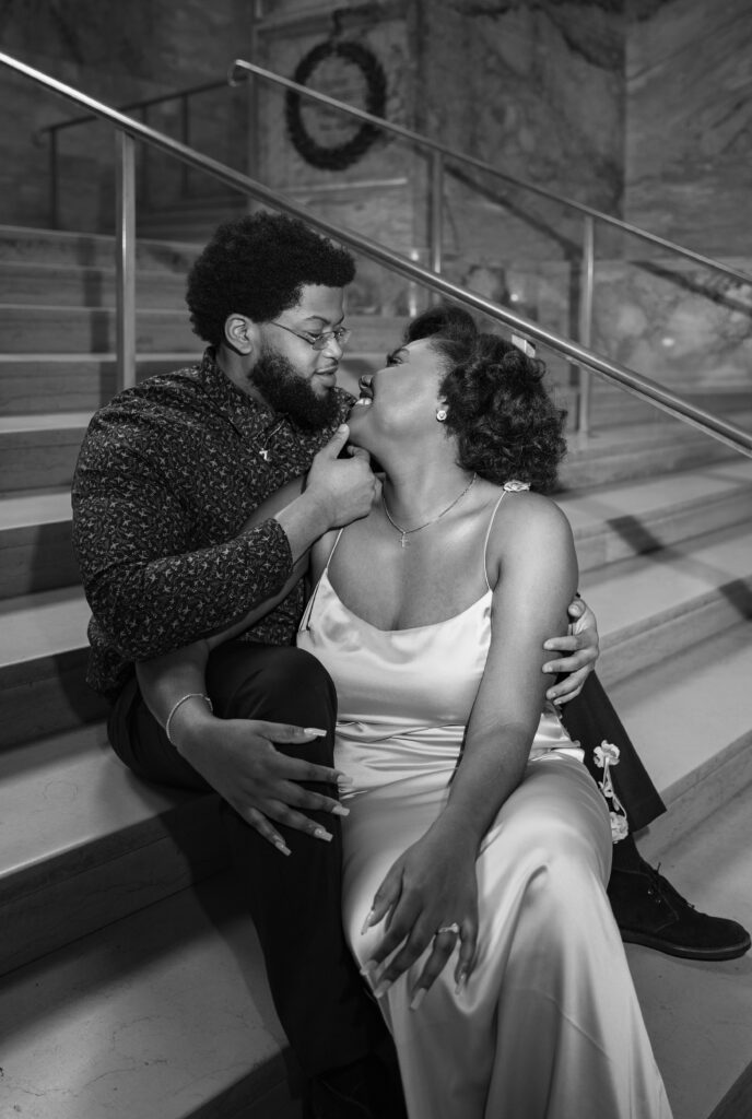 Boston Public Library Engagement Photos Session | Kelsey Sheehan Photography Rhode Island Wedding Photographer | A black and white photo of a couple sits on marble stairs in an embrace. The man gently touches the woman's chin, both smiling warmly at each other, conveying romance and closeness.