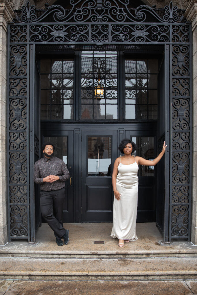 Boston Public Library Engagement Photos Session | Kelsey Sheehan Photography Rhode Island Wedding Photographer | A man and a woman pose confidently in front of an ornate, wrought iron-framed doorway. The woman wears a white satin dress, and the man is in dark attire.