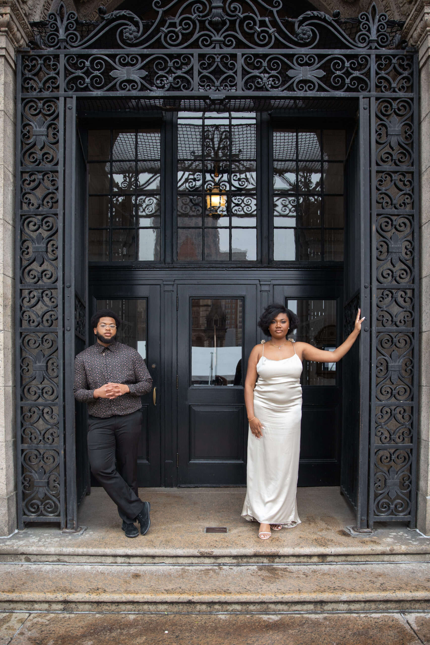 Boston Public Library Engagement Photos Session | Kelsey Sheehan Photography Rhode Island Wedding Photographer | A man and a woman pose confidently in front of an ornate, wrought iron-framed doorway. The woman wears a white satin dress, and the man is in dark attire.