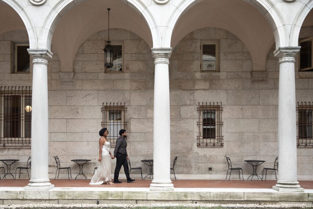 Boston Public Library Engagement Photos Session | Kelsey Sheehan Photography Rhode Island Wedding Photographer | A couple walks side-by-side under an arched, stone colonnade. The woman wears a white dress, the man a dark suit. Patio tables and chairs line the background.