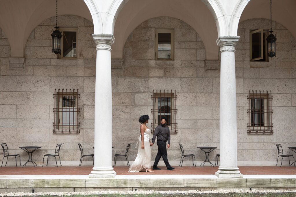 Boston Public Library Engagement Photos Session | Kelsey Sheehan Photography Rhode Island Wedding Photographer | A couple walks hand in hand under elegant stone arches, surrounded by a serene courtyard with tables and chairs, conveying a romantic, peaceful ambiance.