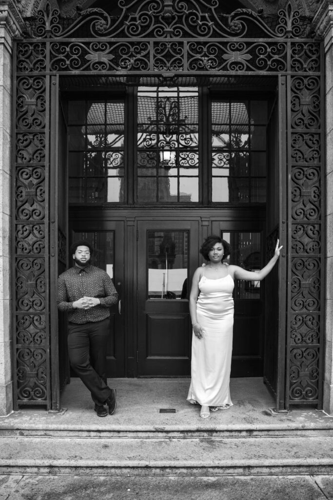 Boston Public Library Engagement Photos Session | Kelsey Sheehan Photography Rhode Island Wedding Photographer | A man and woman stand in ornate, wrought iron doorway. The man is dressed in a dark shirt and pants, the woman in a long, elegant dress. Black and white tones.