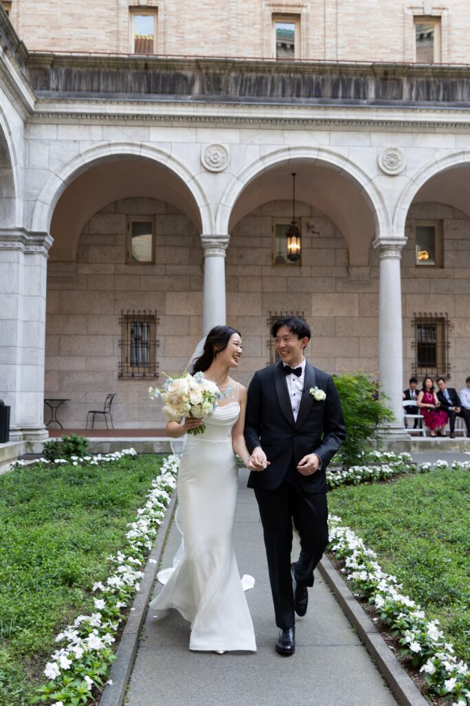 Boston Public Library Wedding | Kelsey Sheehan Photography Rhode Island Wedding Photographer | A joyful couple in wedding attire walks hand in hand down a flower-lined path in a courtyard with arches. They are smiling and share a loving gaze.