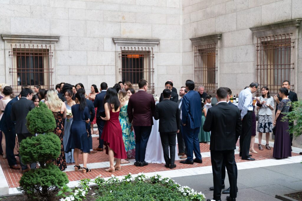 Boston Public Library Wedding | Kelsey Sheehan Photography Rhode Island Wedding Photographer | A crowd of well-dressed people mingles in a courtyard with stone walls and barred windows. The mood is lively and social, with plants in the foreground.