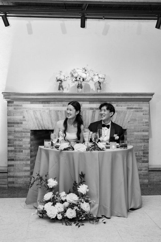 Boston Public Library Wedding | Kelsey Sheehan Photography Rhode Island Wedding Photographer | A joyful couple sits at a round table adorned with flowers, in front of a brick fireplace. Both are smiling warmly, creating a festive and loving atmosphere.
