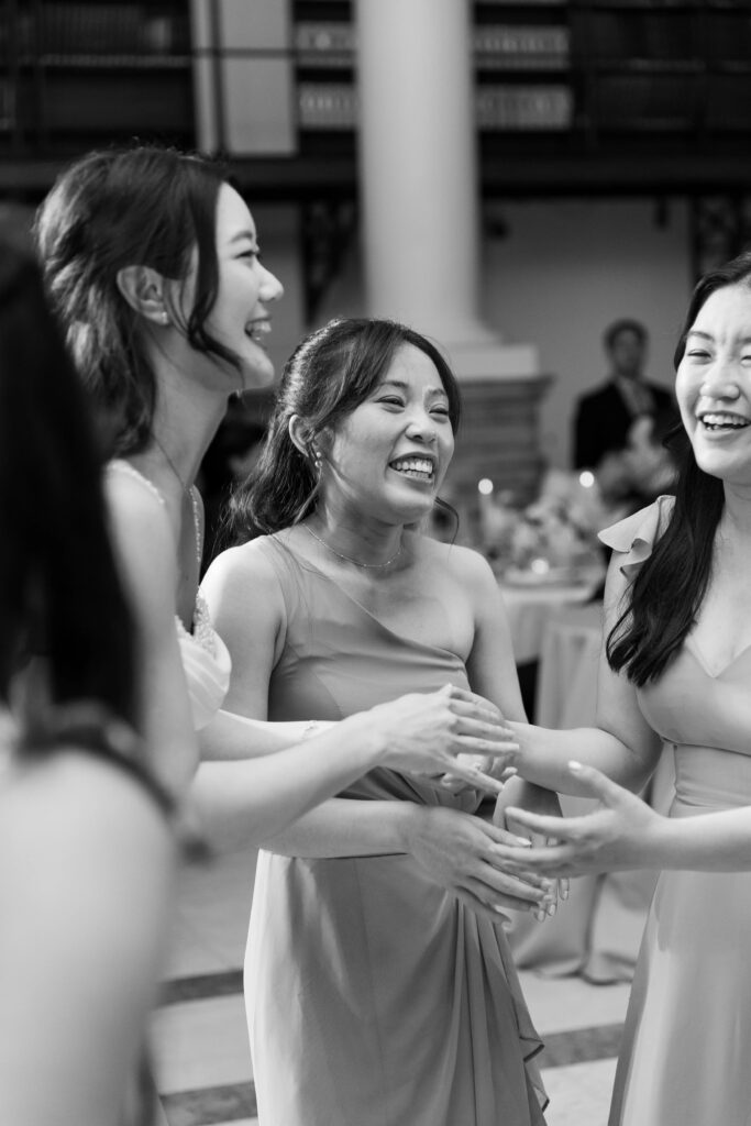 Boston Public Library Wedding | Kelsey Sheehan Photography Rhode Island Wedding Photographer | Three women in elegant dresses are laughing and holding hands at an indoor event. They convey joy and connection in this black-and-white photo.