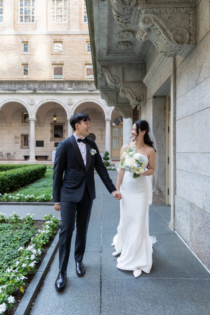 Boston Public Library Wedding | Kelsey Sheehan Photography Rhode Island Wedding Photographer | A bride and groom walk hand in hand, smiling at each other in an elegant courtyard. The bride holds a bouquet, and the groom wears a black tuxedo.