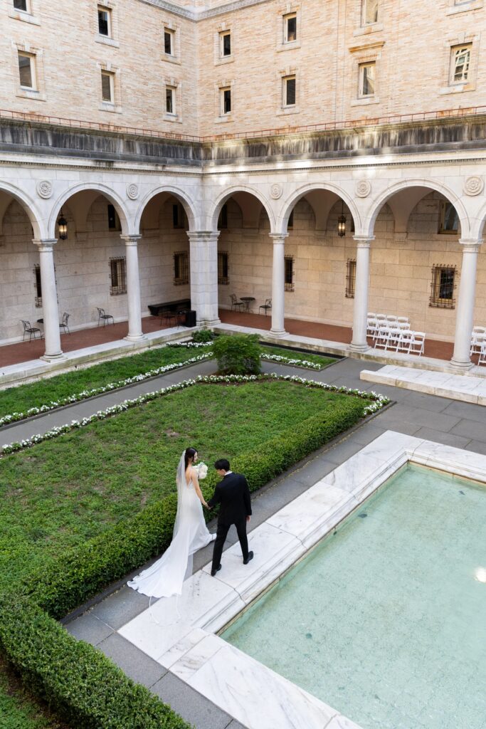 Boston Public Library Wedding | Kelsey Sheehan Photography Rhode Island Wedding Photographer | A bride and groom walk hand in hand in a historic courtyard with arches. A serene pool and green hedges surround them, evoking a romantic atmosphere.