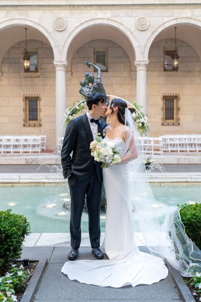 Boston Public Library Wedding | Kelsey Sheehan Photography Rhode Island Wedding Photographer | Bride and groom kiss before a fountain, with the bride in a white gown and veil holding a bouquet, set against an elegant arched courtyard backdrop.