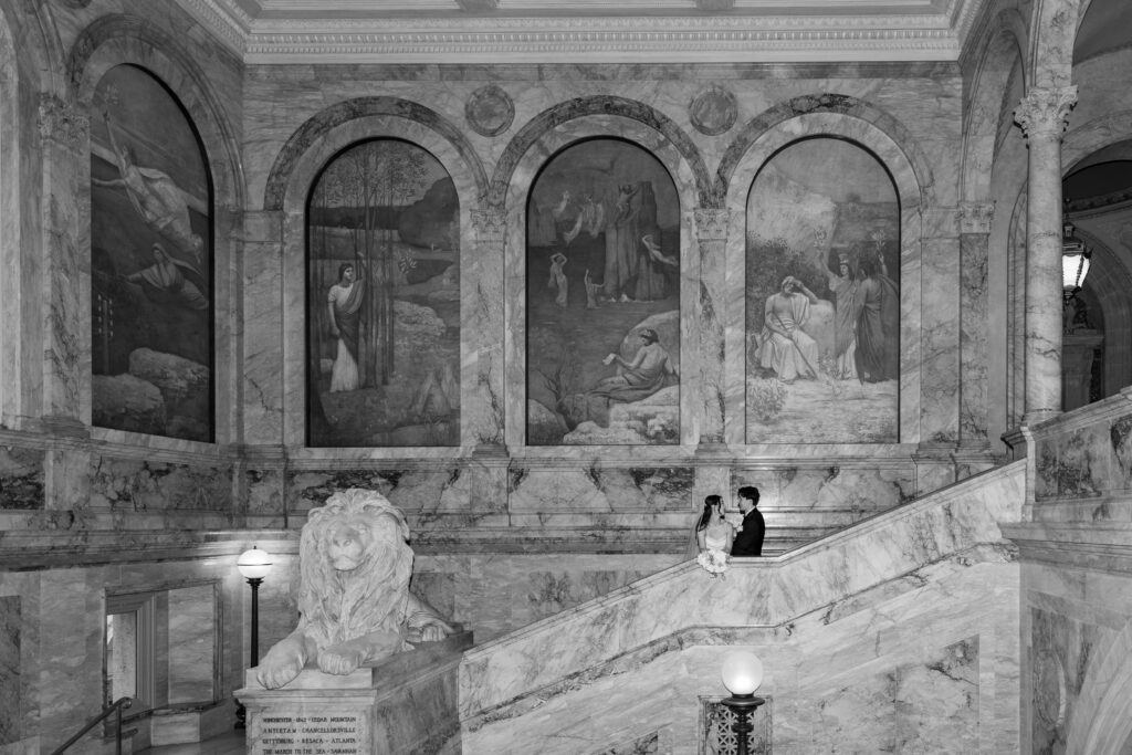 Boston Public Library Wedding | Kelsey Sheehan Photography Rhode Island Wedding Photographer | A couple stands on a grand marble staircase in an ornate, historical building. Behind them are three large frescoes in arched frames. A stone lion statue adds elegance to the scene. The image conveys romance and timeless beauty.