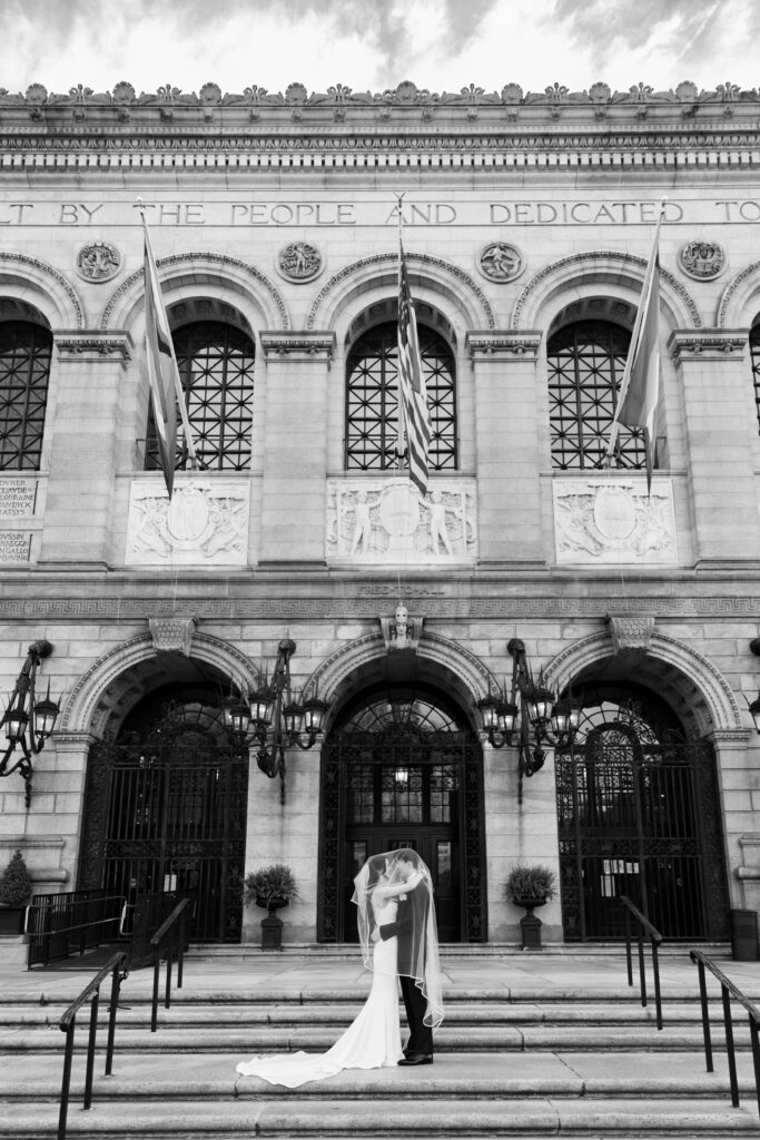 Boston Public Library Wedding | Kelsey Sheehan Photography Rhode Island Wedding Photographer | A bride and groom share a kiss under a veil on the steps of a historic, ornate building with arched windows and flags, conveying romance and elegance.