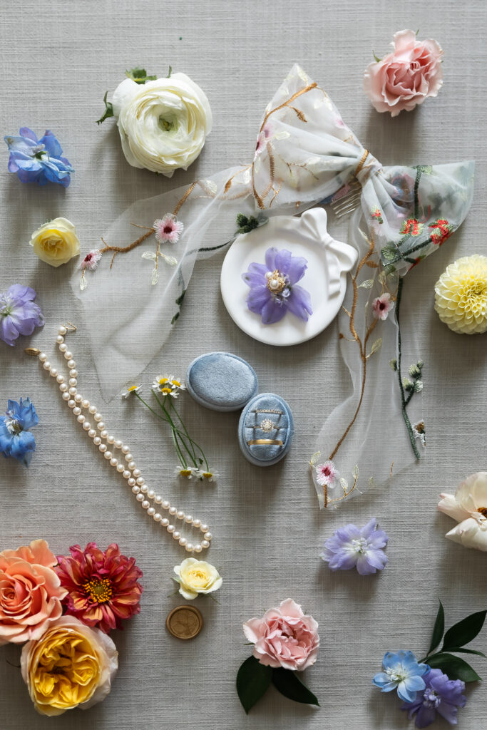 Bristol Harbor Inn | Kelsey Sheehan Photography Rhode Island Wedding Photographer | A flatlay of colorful flowers, a pearl necklace, blue ring boxes, sheer embroidered fabric, and a delicate dish on a neutral background.