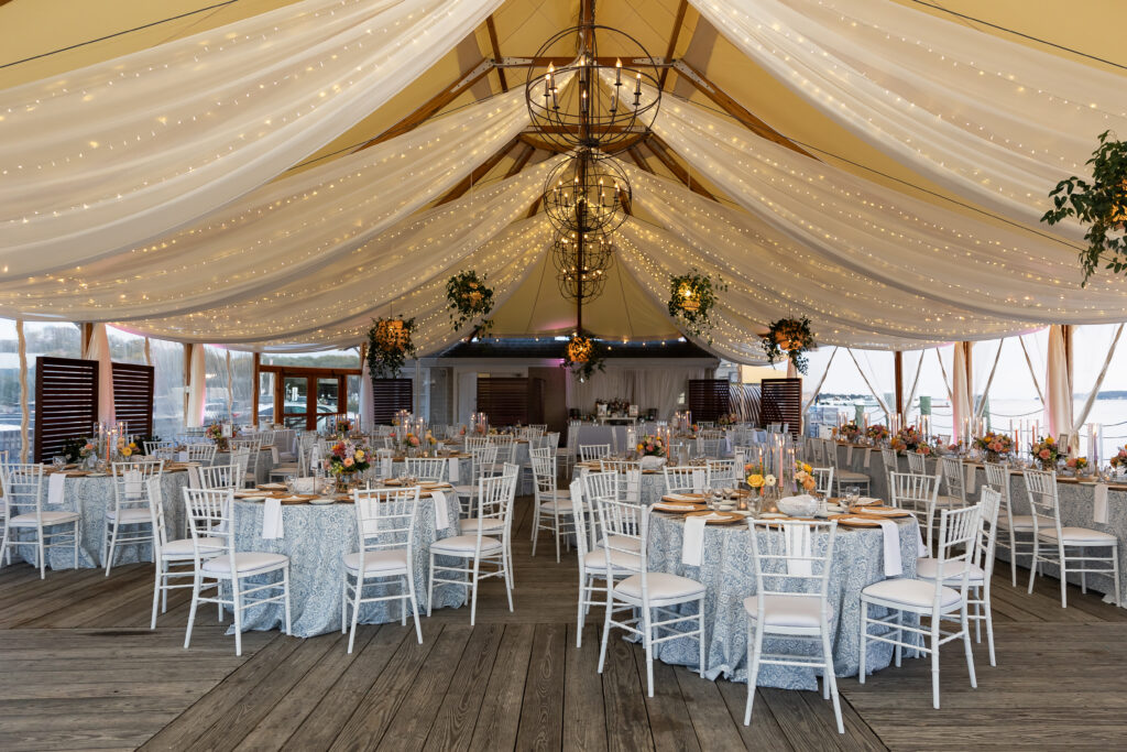 Bristol Harbor Inn | Kelsey Sheehan Photography Rhode Island Wedding Photographer | Elegant wedding reception inside a tent with draped fabric and string lights. Round tables with floral centerpieces and white chairs create a festive ambiance.