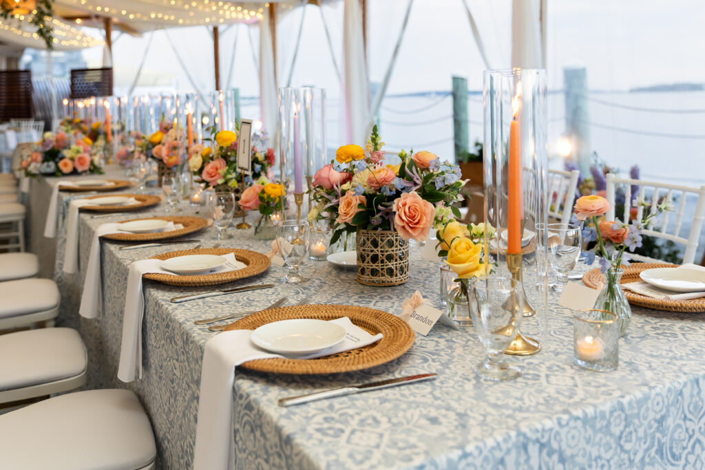 Bristol Harbor Inn | Kelsey Sheehan Photography Rhode Island Wedding Photographer | Elegant wedding table setting with floral centerpieces, soft candlelight, and blue patterned tablecloths beside a serene waterside view.