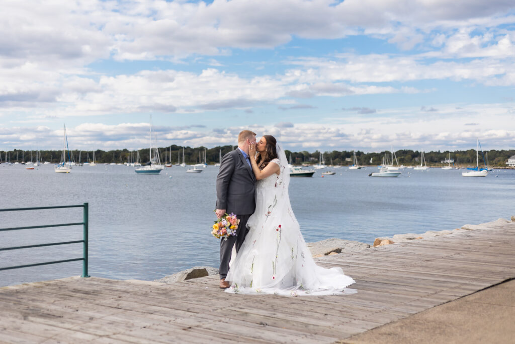 Bristol Harbor Inn | Kelsey Sheehan Photography Rhode Island Wedding Photographer | A couple kisses on a wooden pier by a tranquil lake. The bride wears a floral gown, holding colorful flowers. Sailboats dot the water under a partly cloudy sky. Romance and joy fill the scene.