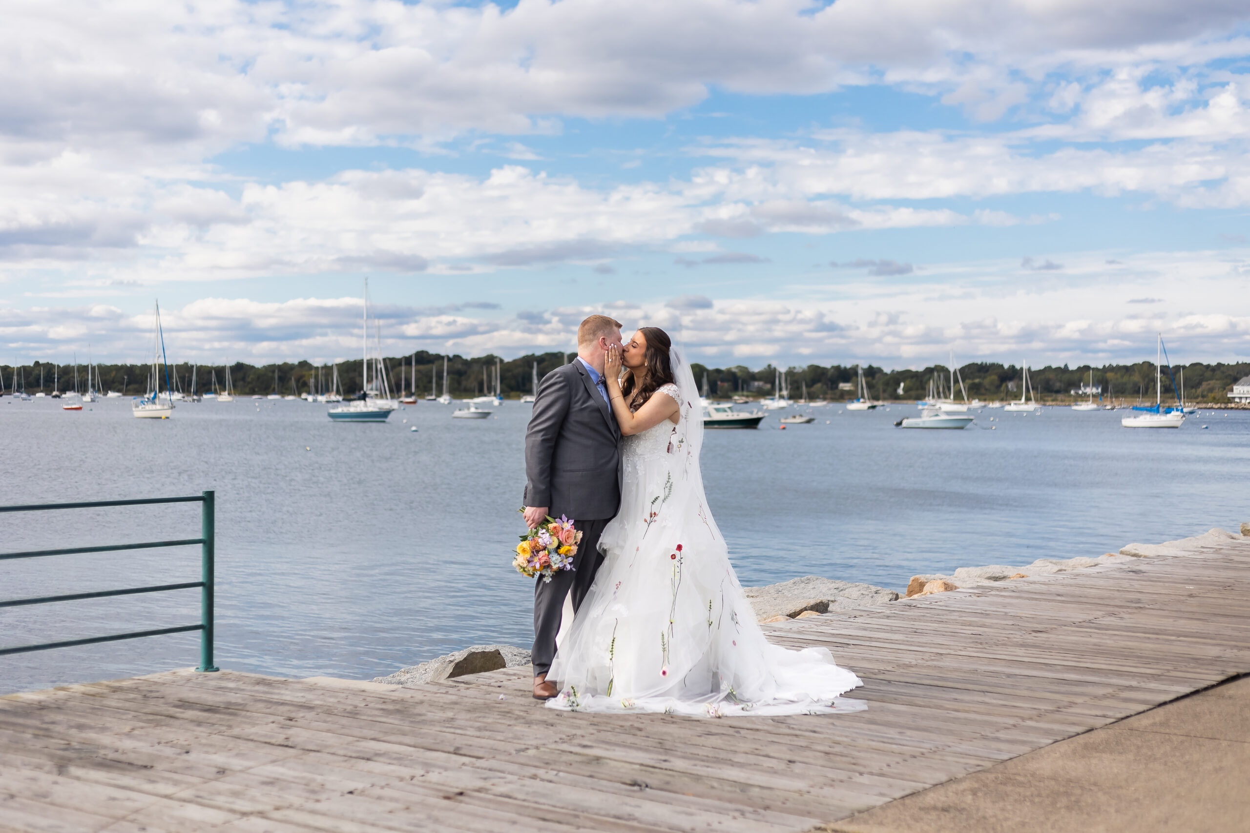 Bristol Harbor Inn | Kelsey Sheehan Photography Rhode Island Wedding Photographer | A couple kisses on a wooden pier by a tranquil lake. The bride wears a floral gown, holding colorful flowers. Sailboats dot the water under a partly cloudy sky. Romance and joy fill the scene.