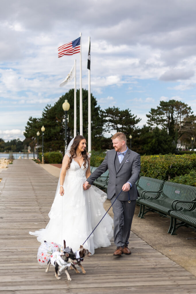 Bristol Harbor Inn | Kelsey Sheehan Photography Rhode Island Wedding Photographer | Bride and groom smiling, walking on a boardwalk with two small dogs in bridal attire. Flags and trees in the background under a cloudy sky.