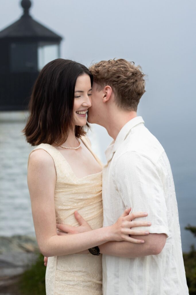 Castle Hill Inn | Kelsey Sheehan Photography Rhode Island Wedding Photographer | A couple embraces by a misty lighthouse. The woman smiles warmly while the man whispers. The mood is intimate and serene, with an overcast sky.
