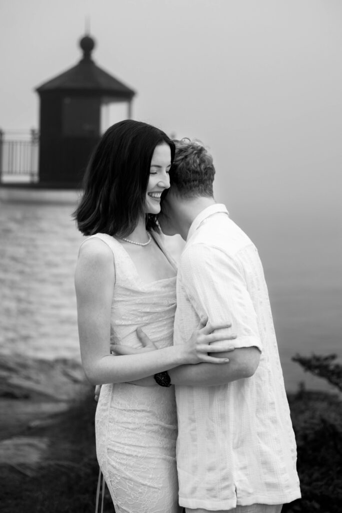 Castle Hill Inn | Kelsey Sheehan Photography Rhode Island Wedding Photographer | Black and white photo of a smiling couple embracing near a foggy shoreline with a lighthouse in the background. The scene feels romantic and serene.