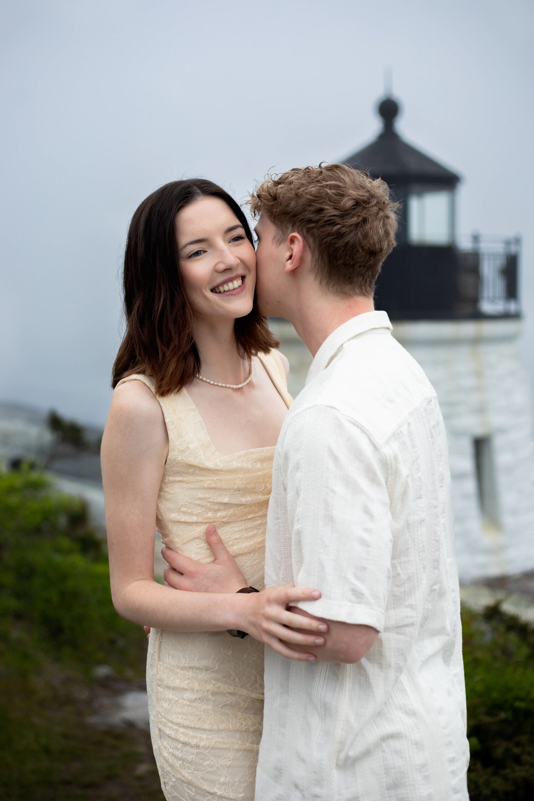 Castle Hill Inn | Kelsey Sheehan Photography Rhode Island Wedding Photographer | A smiling woman in a light dress embraces a man in a white shirt near a lighthouse. The scene conveys warmth and happiness, set against a misty background.