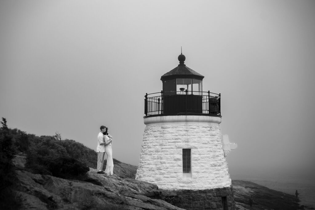 Castle Hill Inn | Kelsey Sheehan Photography Rhode Island Wedding Photographer | A couple embraces on a rocky cliff near a lighthouse in a foggy setting. The black-and-white image conveys a romantic and serene atmosphere.