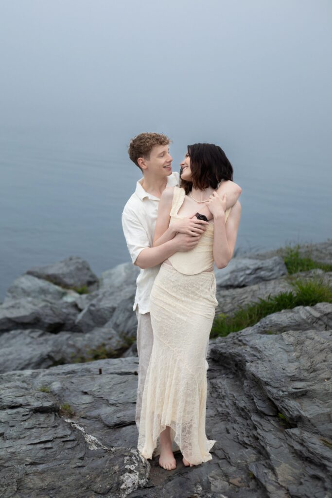Castle Hill Inn | Kelsey Sheehan Photography Rhode Island Wedding Photographer | A couple embraces on rocky terrain by a calm, misty lake. The woman in a cream dress smiles at the man in a white shirt, conveying a peaceful, romantic mood.