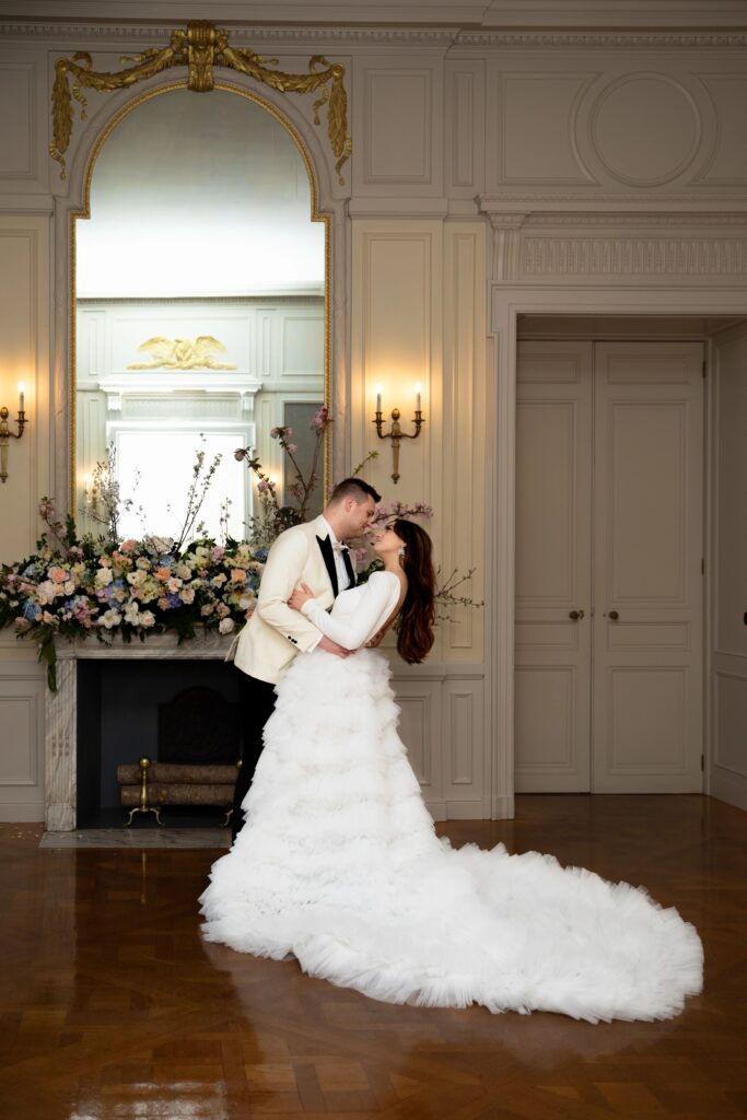Glen Manor House | Kelsey Sheehan Photography Rhode Island Wedding Photographer | A couple shares a romantic embrace in an elegant room. She wears a white, ruffled gown; he wears a white jacket. A floral arrangement and large mirror are behind them.