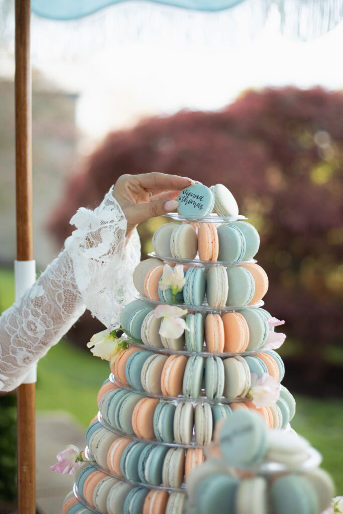 Glen Manor House | Kelsey Sheehan Photography Rhode Island Wedding Photographer | A hand in lace places a macaron atop a pastel macaron tower adorned with small flowers. The scene feels elegant and celebratory in an outdoor setting.