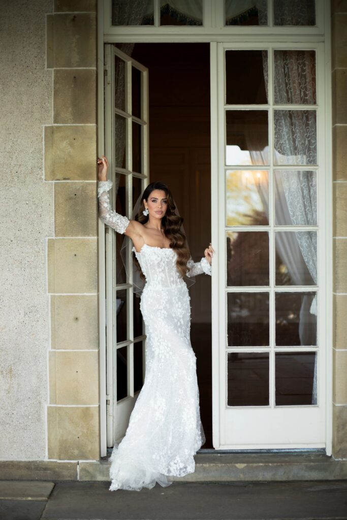 Castle Wedding Venues in New England | Kelsey Sheehan Photography Rhode Island Wedding Photographer | A woman in a long, elegant white gown with lace details stands confidently in a doorway, framed by large glass doors and neutral-toned walls.