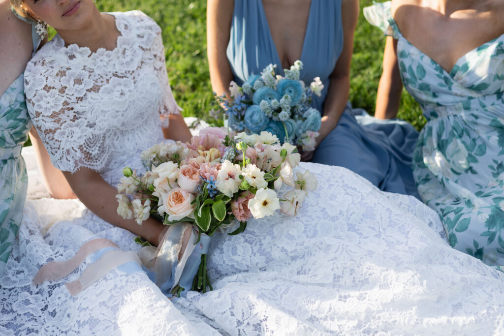 Glen Manor House | Kelsey Sheehan Photography Rhode Island Wedding Photographer | Three women in elegant dresses sit on grass, holding floral bouquets. The central bouquet features pastel roses and greenery, exuding a romantic tone.