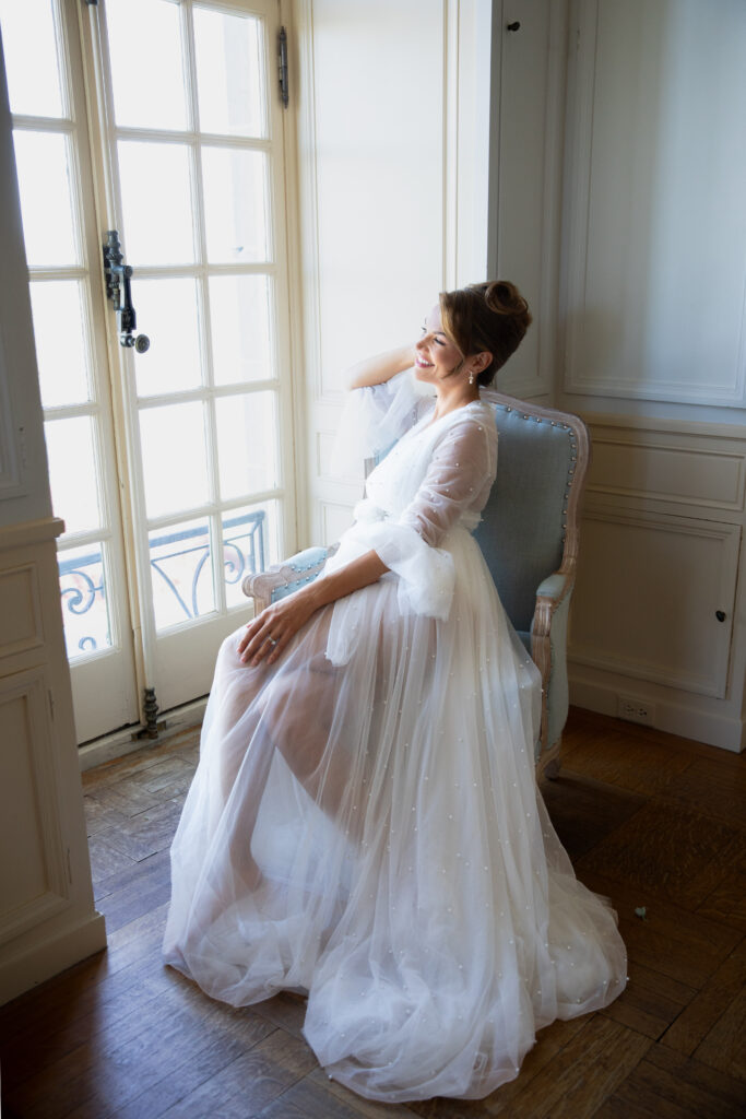 Glen Manor House | Kelsey Sheehan Photography Rhode Island Wedding Photographer | Image Woman in a flowing white robe sits on a chair, smiling softly at sunlight streaming through French doors. The scene conveys elegance and tranquility.