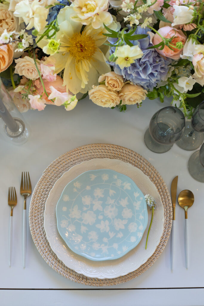 Bristol Harbor Inn | Kelsey Sheehan Photography Rhode Island Wedding Photographer | Elegant table setting with a floral arrangement. A light blue plate with white flowers rests on woven placemat, surrounded by gold cutlery and delicate blossoms.